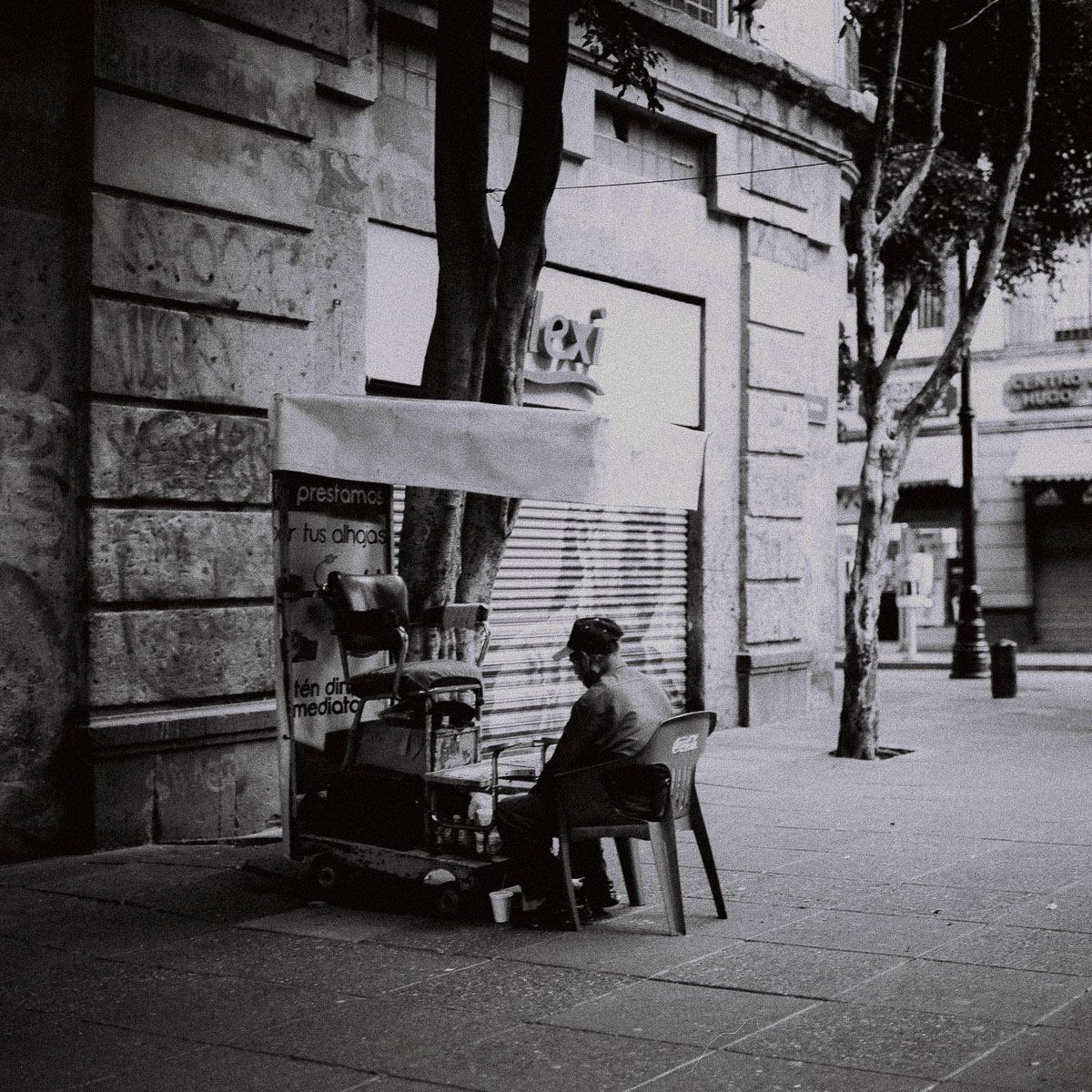 Black-and-white film photograph of a seated street vendor on a Mexico City sidewalk.