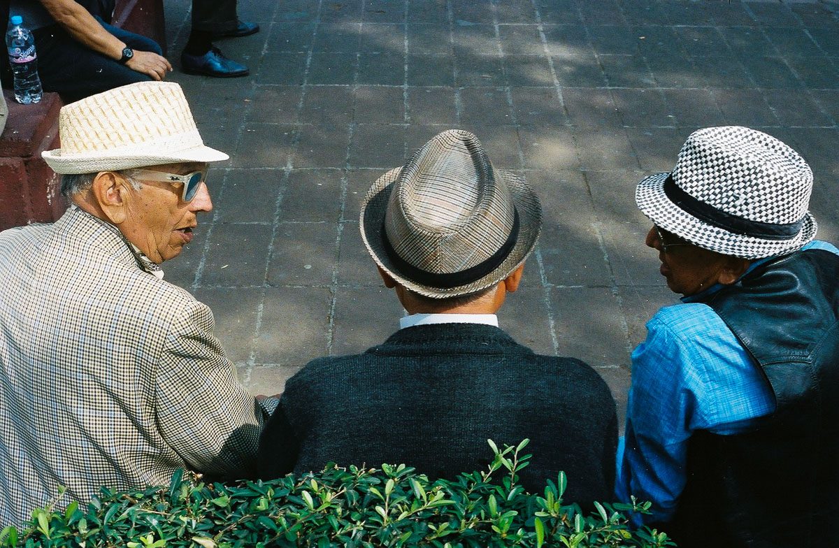 Color analog street photograph of three older men in patterned hats talking on a bench.