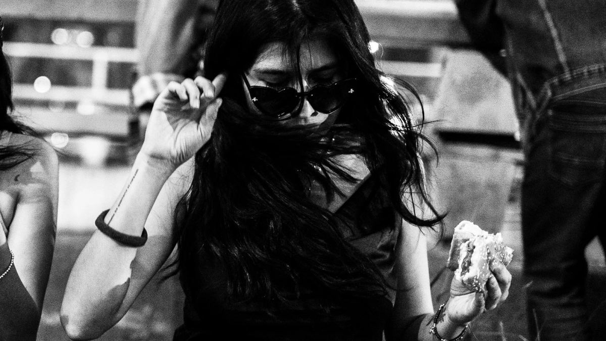 Black-and-white photograph of a woman holding a burger while adjusting her sunglasses