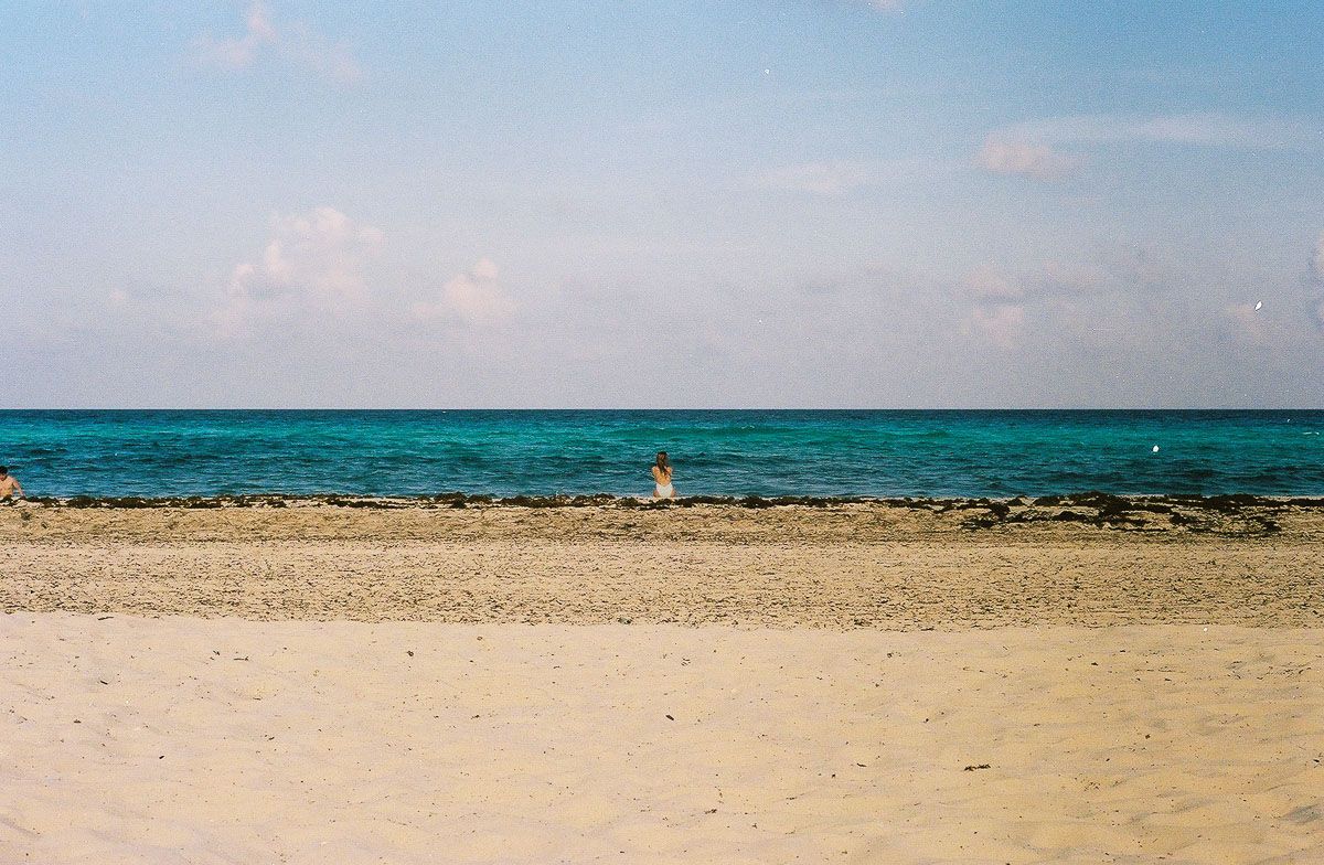 Color film photograph of a distant beachgoer standing at the turquoise Caribbean shoreline beneath a pale sky.
