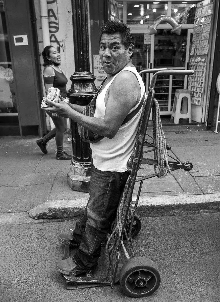 Black-and-white street portrait of a man standing beside a hand truck