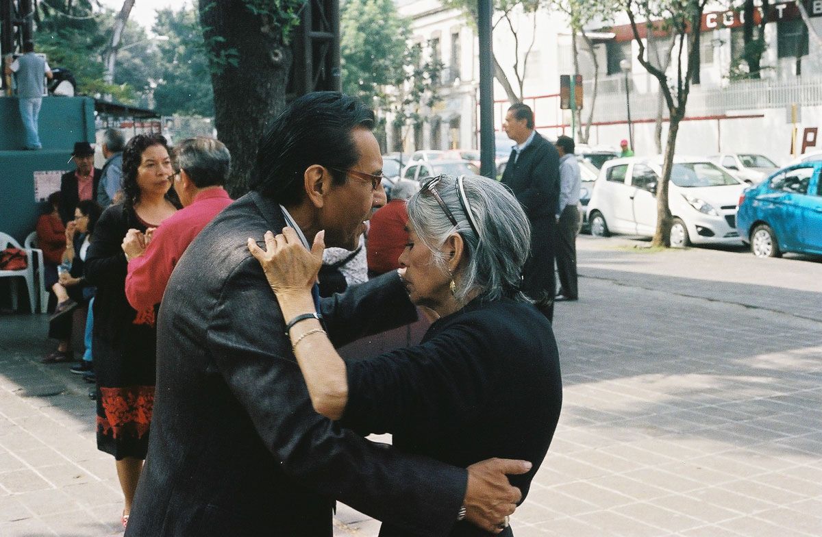 Color film photograph of an older couple embracing while dancing in the street.