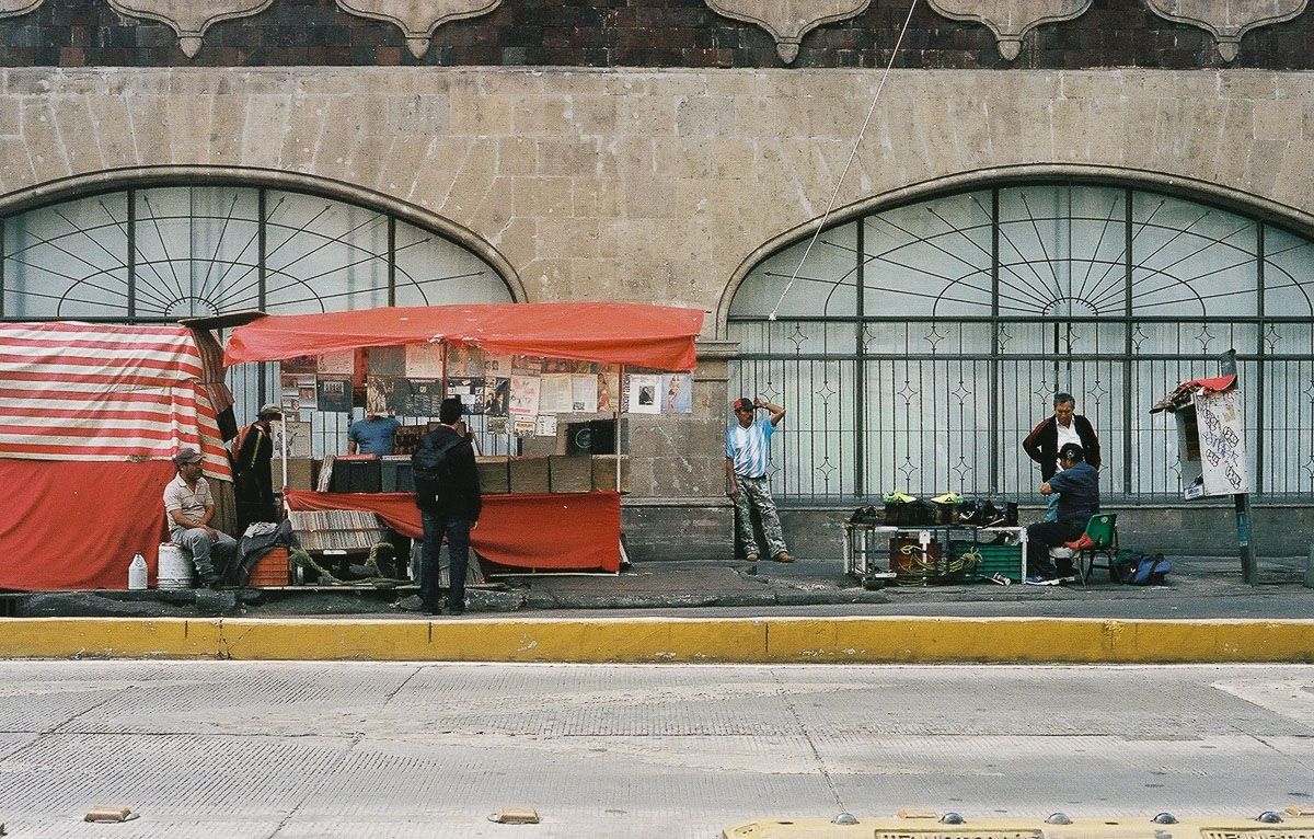 Color film photograph of street vendors beneath red market awnings in Mexico City.