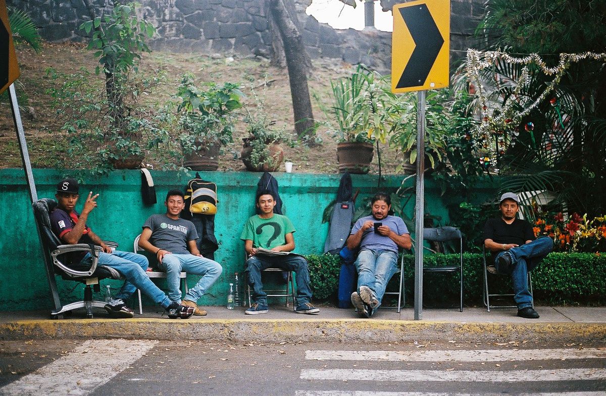 Color film street photograph of five men seated together beside a green wall.