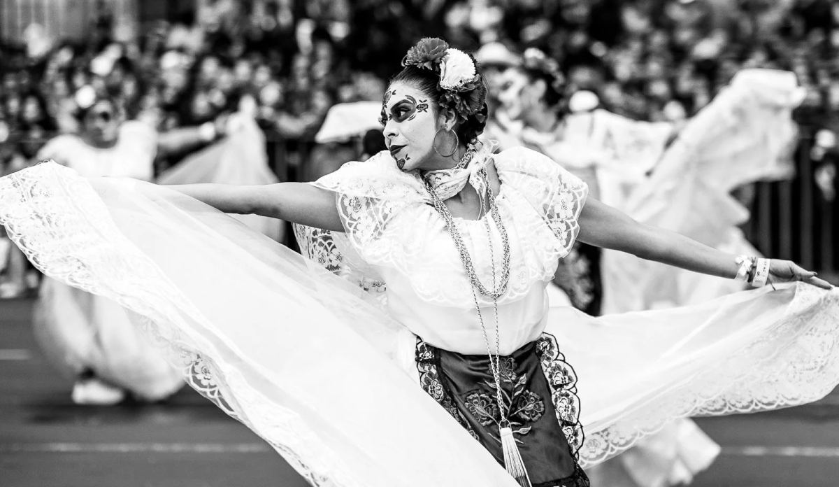 Dancer with skull face paint spreading a white cape during a Day of the Dead parade
