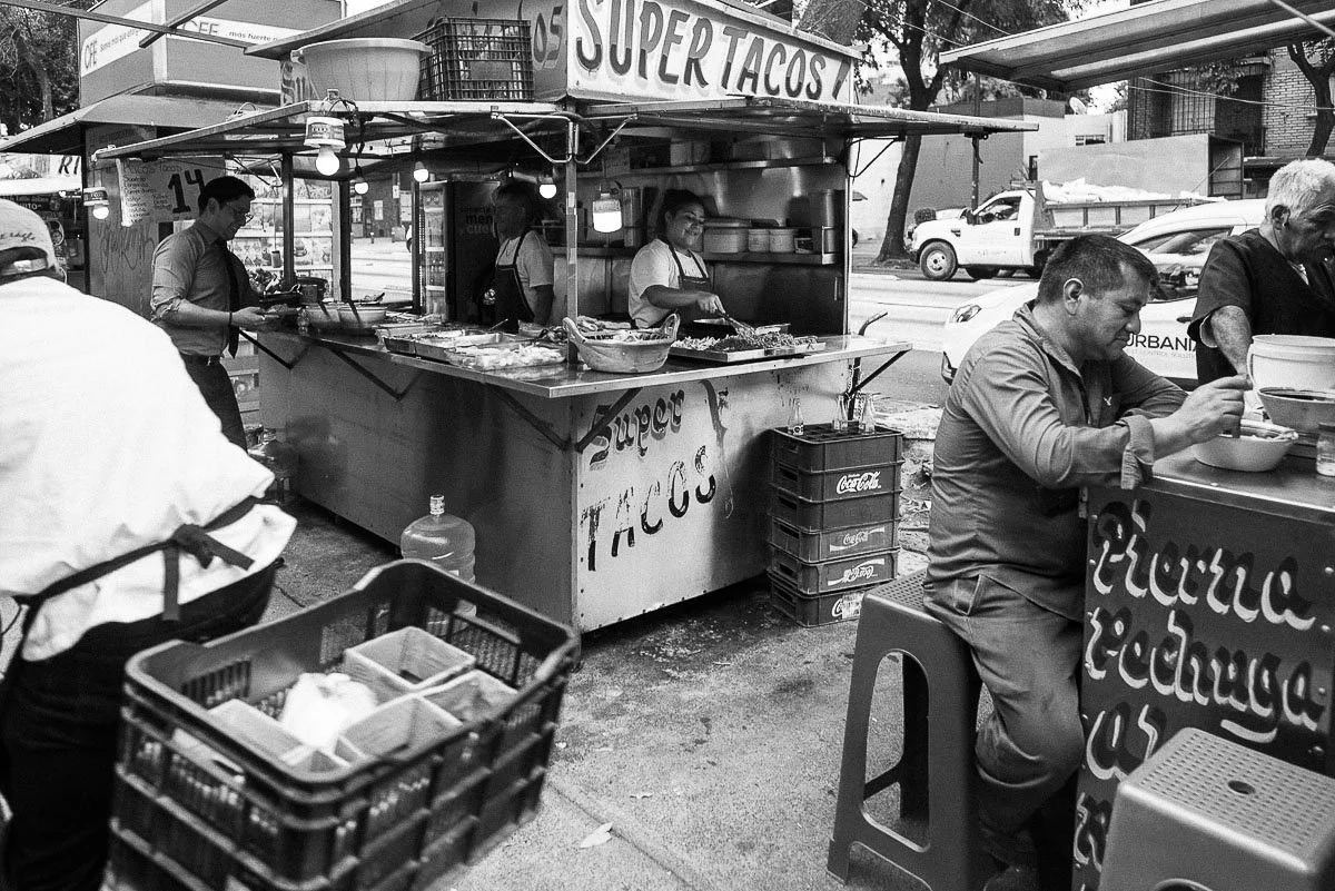 Black-and-white street photograph of customers eating at a Super Tacos stand