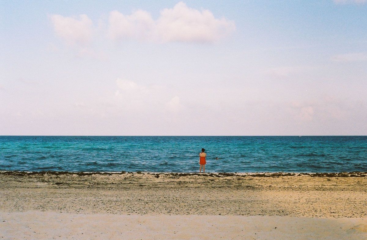Color analog photograph of a woman in red standing at the edge of the ocean.