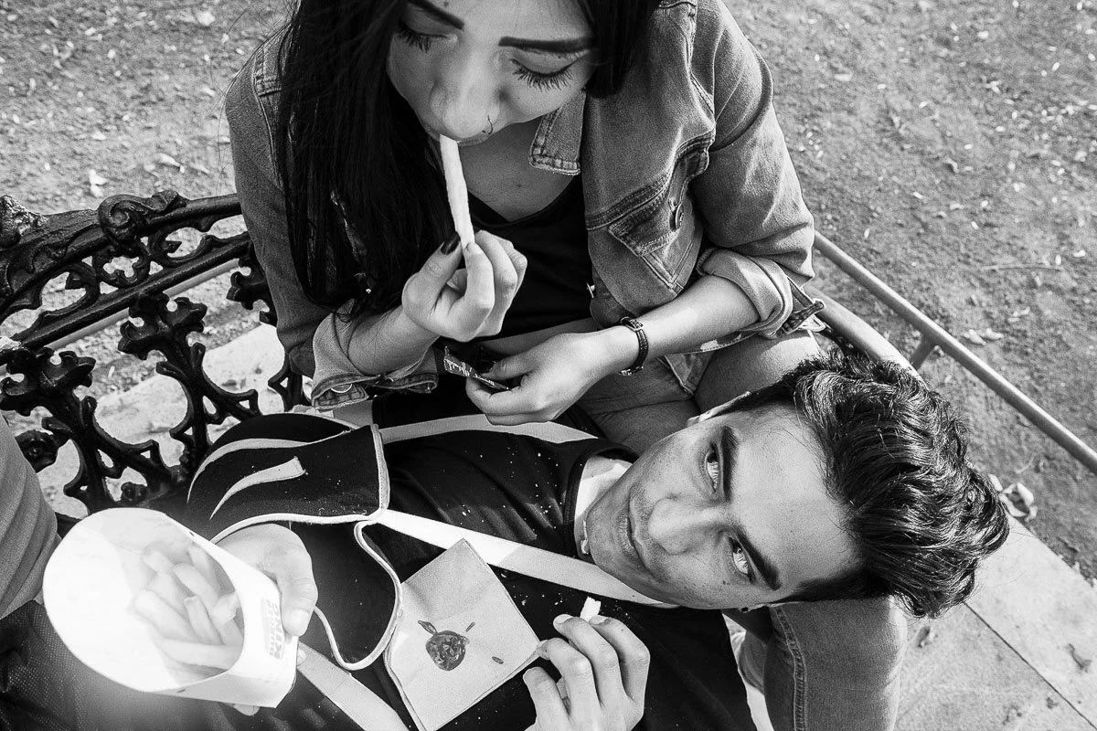 Overhead black-and-white photograph of a woman eating fries beside a man resting on a park bench