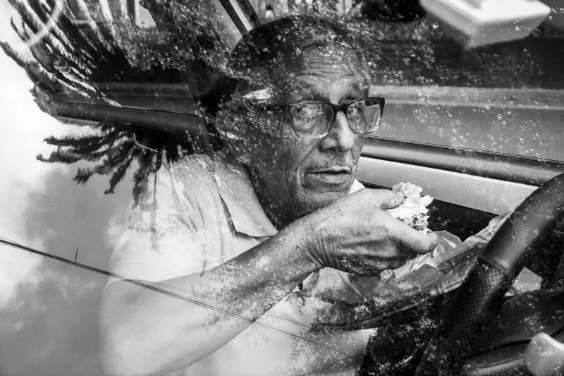 Black-and-white photograph of a man with dreadlocks eating a burger inside a car