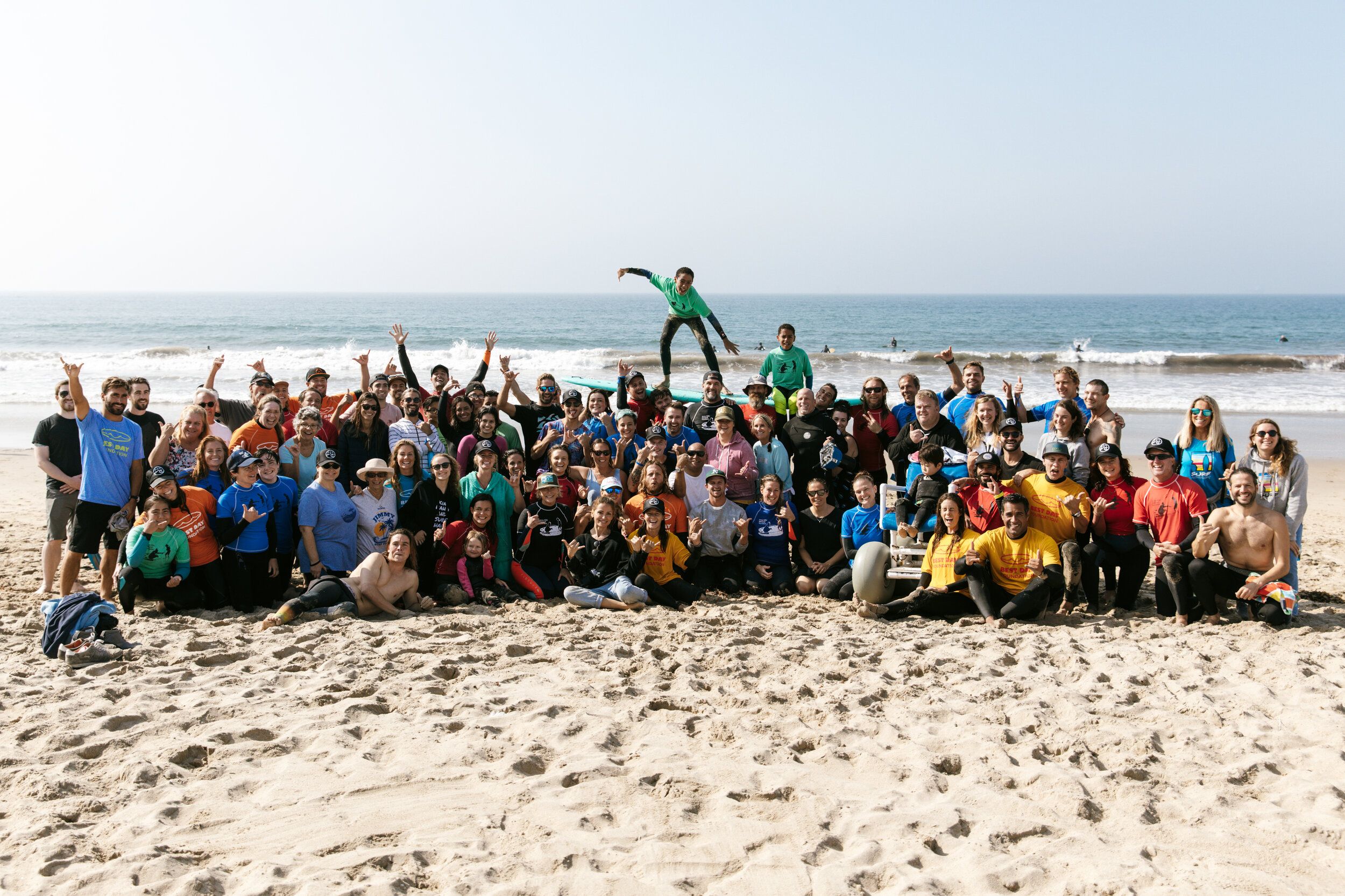 Large group photo of surf therapy volunteers and participants on the beach, with a boy leaping in the center