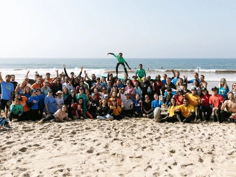 Large group photo of surf therapy volunteers and participants on the beach, with a boy leaping in the center