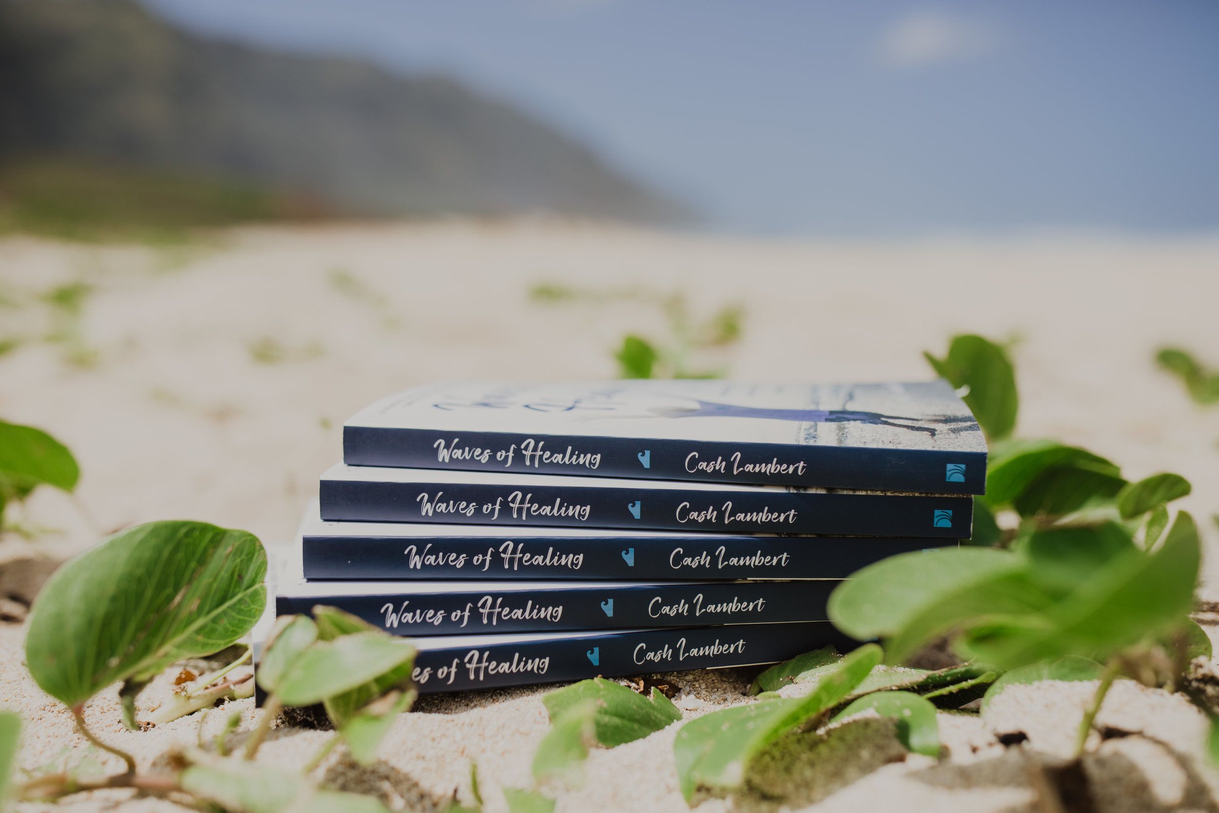A stack of Waves of Healing paperbacks on the sand among beach vines