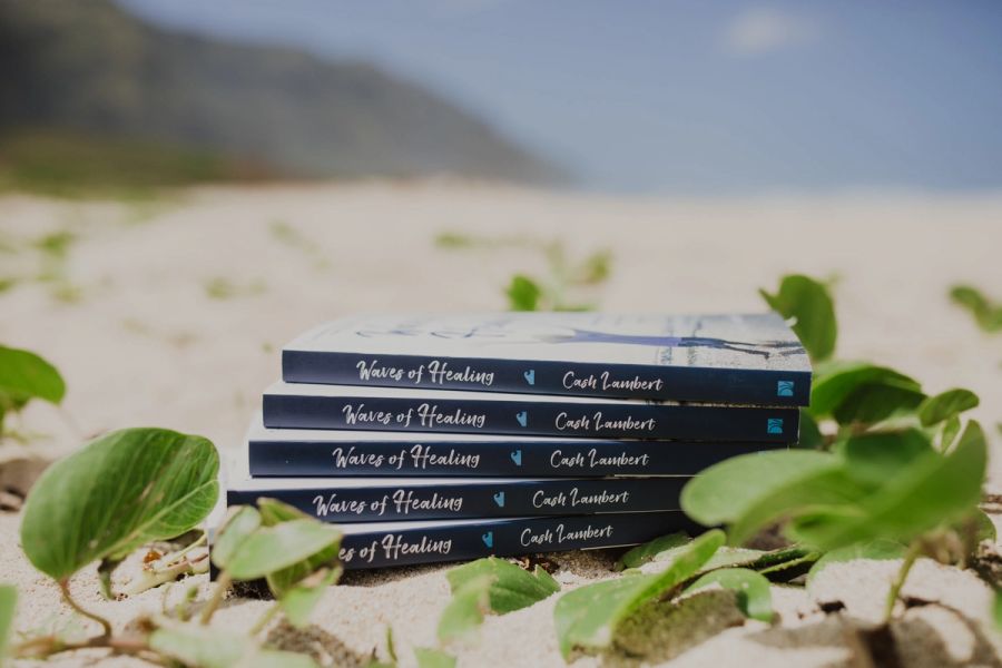 A stack of Waves of Healing paperbacks on the sand among beach vines