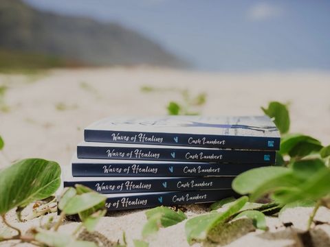 A stack of Waves of Healing paperbacks on the sand among beach vines