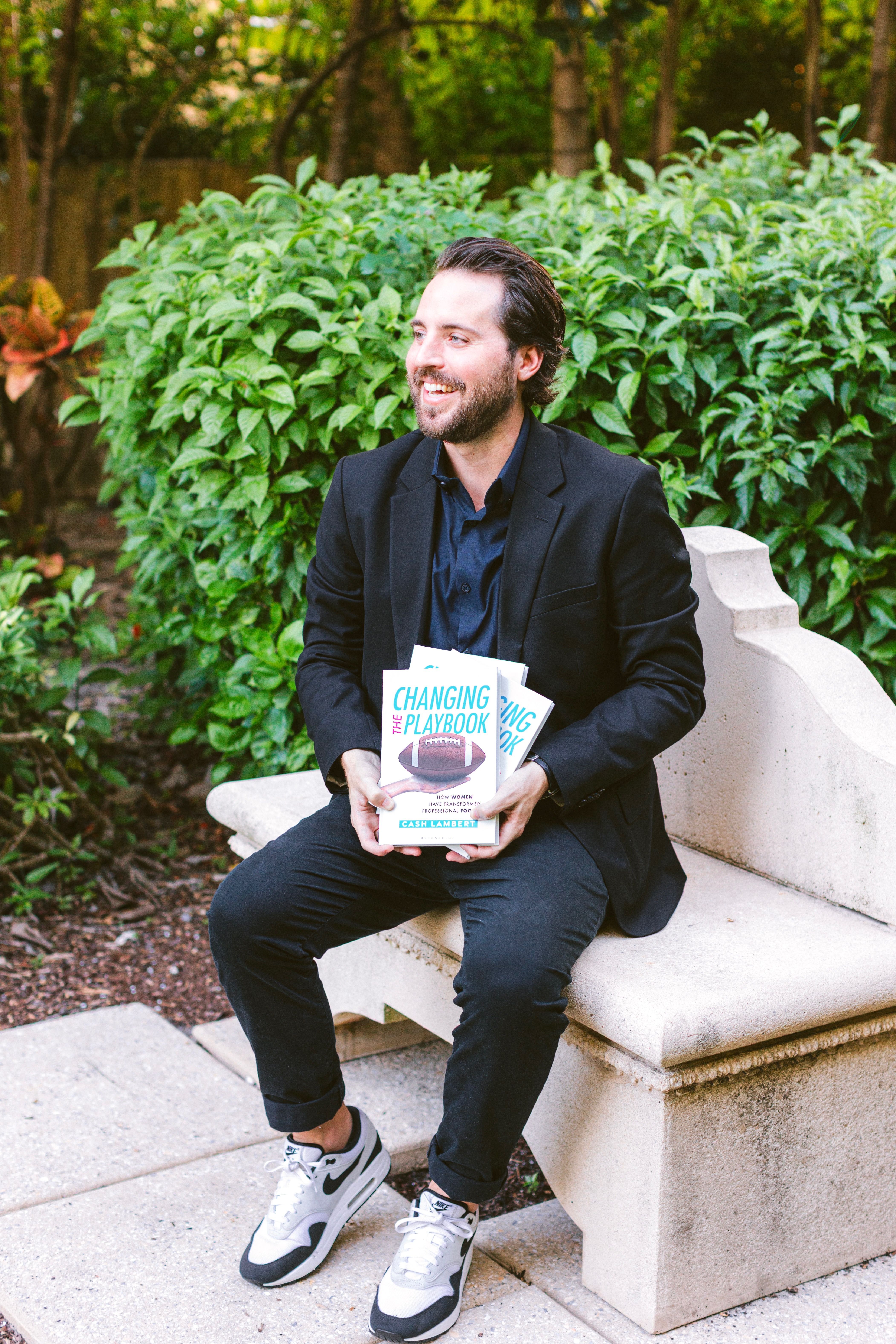 Cash Lambert on a stone bench, smiling, holding copies of Changing the Playbook