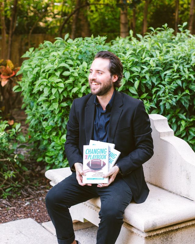 Cash Lambert on a stone bench, smiling, holding copies of Changing the Playbook