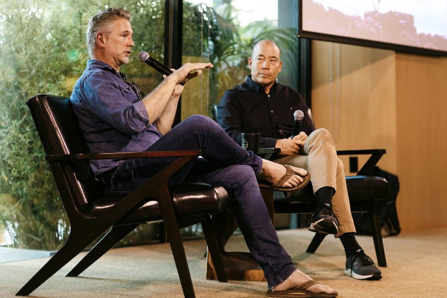 Two men in a panel discussion, one holding a microphone and gesturing, beside a screen showing video
