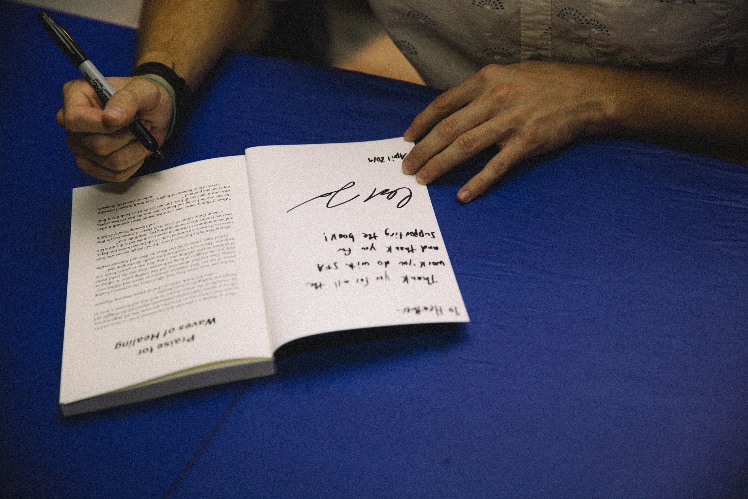 Close-up of hands signing a copy of Waves of Healing on a blue tablecloth