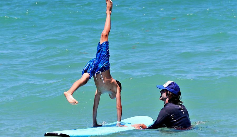 A boy performing a handstand on a surfboard while an instructor holds it steady in the waves
