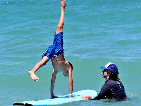 A boy performing a handstand on a surfboard while an instructor holds it steady in the waves