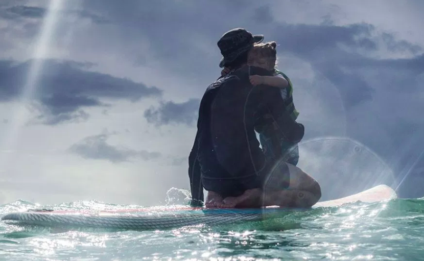 A man in a wetsuit hugging a child on a paddleboard, silhouetted against a cloudy sky
