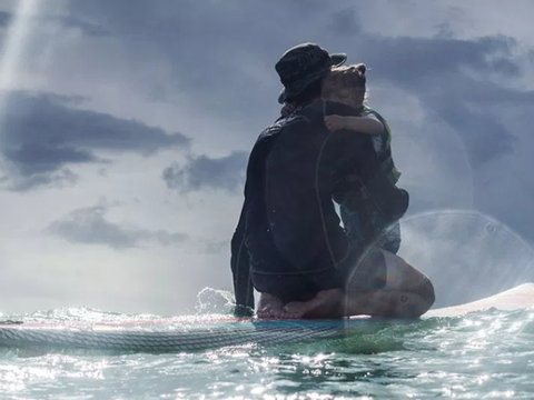 A man in a wetsuit hugging a child on a paddleboard, silhouetted against a cloudy sky