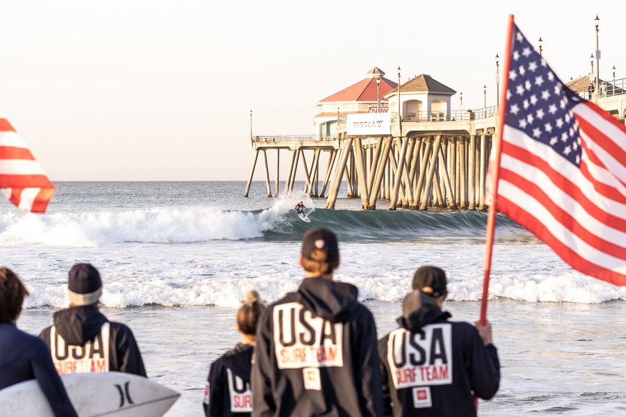 A surfer riding a wave near the Huntington Beach pier as USA Surf Team members watch from shore
