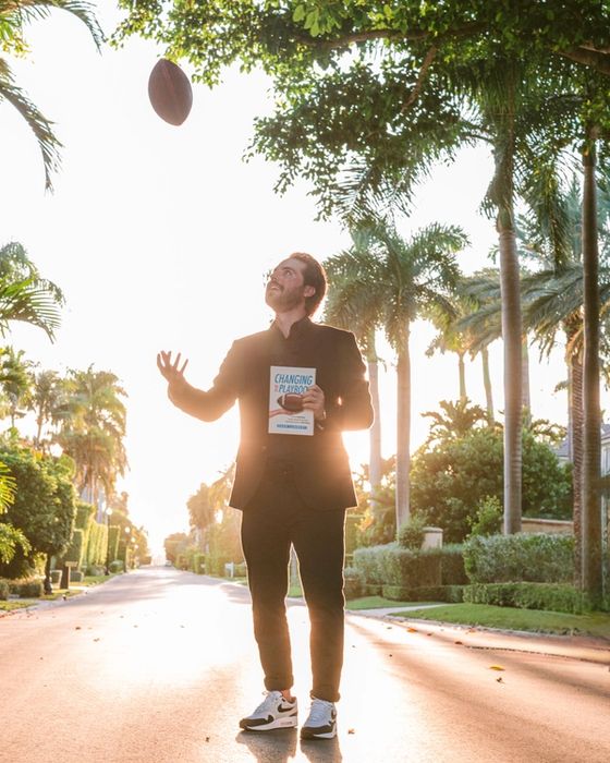Cash Lambert on a palm-lined street, tossing a football and holding a copy of Changing the Playbook