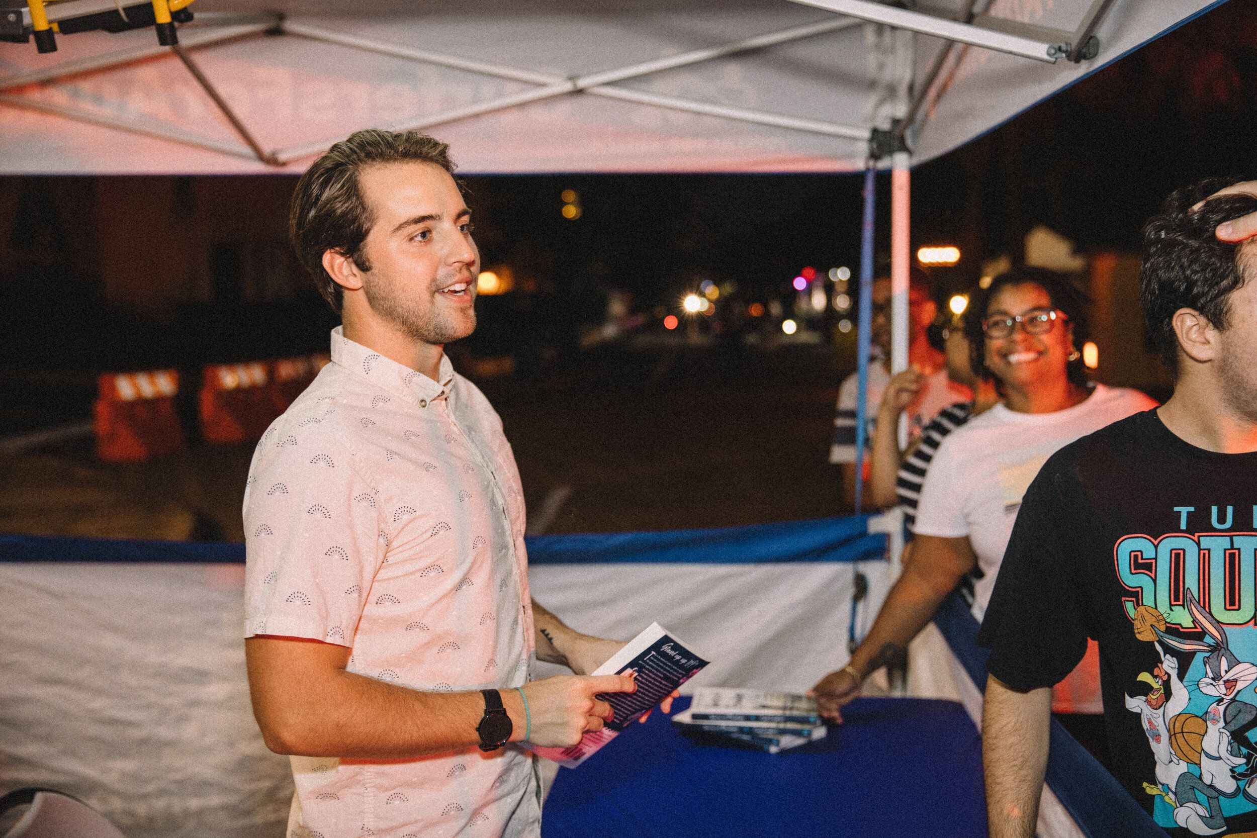Cash Lambert smiling while holding a book and flyer at a nighttime outdoor booth