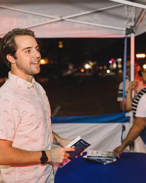 Cash Lambert smiling while holding a book and flyer at a nighttime outdoor booth