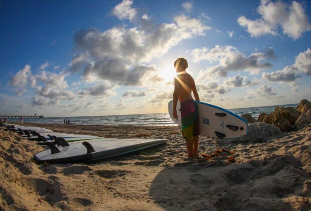 Boy holding a surfboard and silhouetted by the sun, standing on the beach beside a row of surfboards