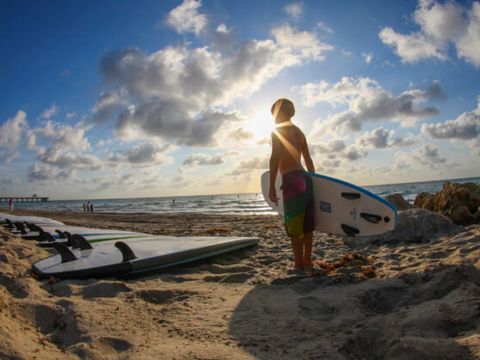 Boy holding a surfboard and silhouetted by the sun, standing on the beach beside a row of surfboards