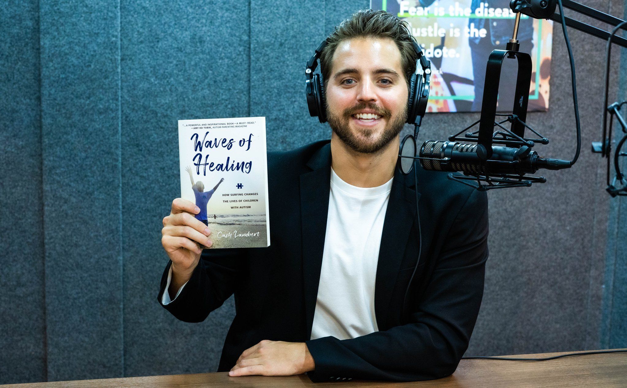 Cash Lambert wearing headphones and holding Waves of Healing in front of a podcast microphone
