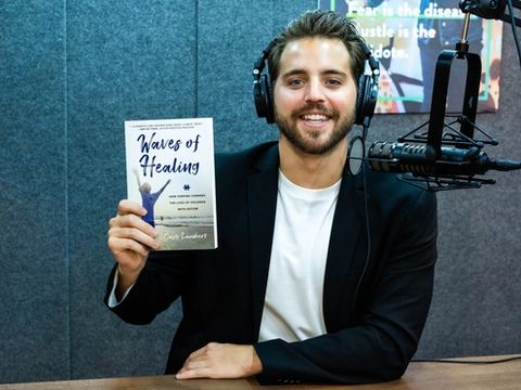 Cash Lambert wearing headphones and holding Waves of Healing in front of a podcast microphone