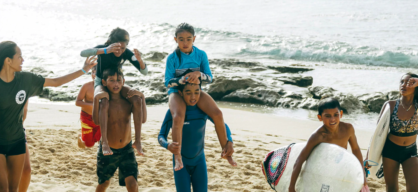 Children giving each other piggyback rides on the beach near the water, with surfboards nearby