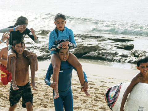 Children giving each other piggyback rides on the beach near the water, with surfboards nearby