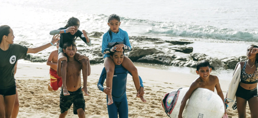 Children giving each other piggyback rides on the beach near the water, with surfboards nearby