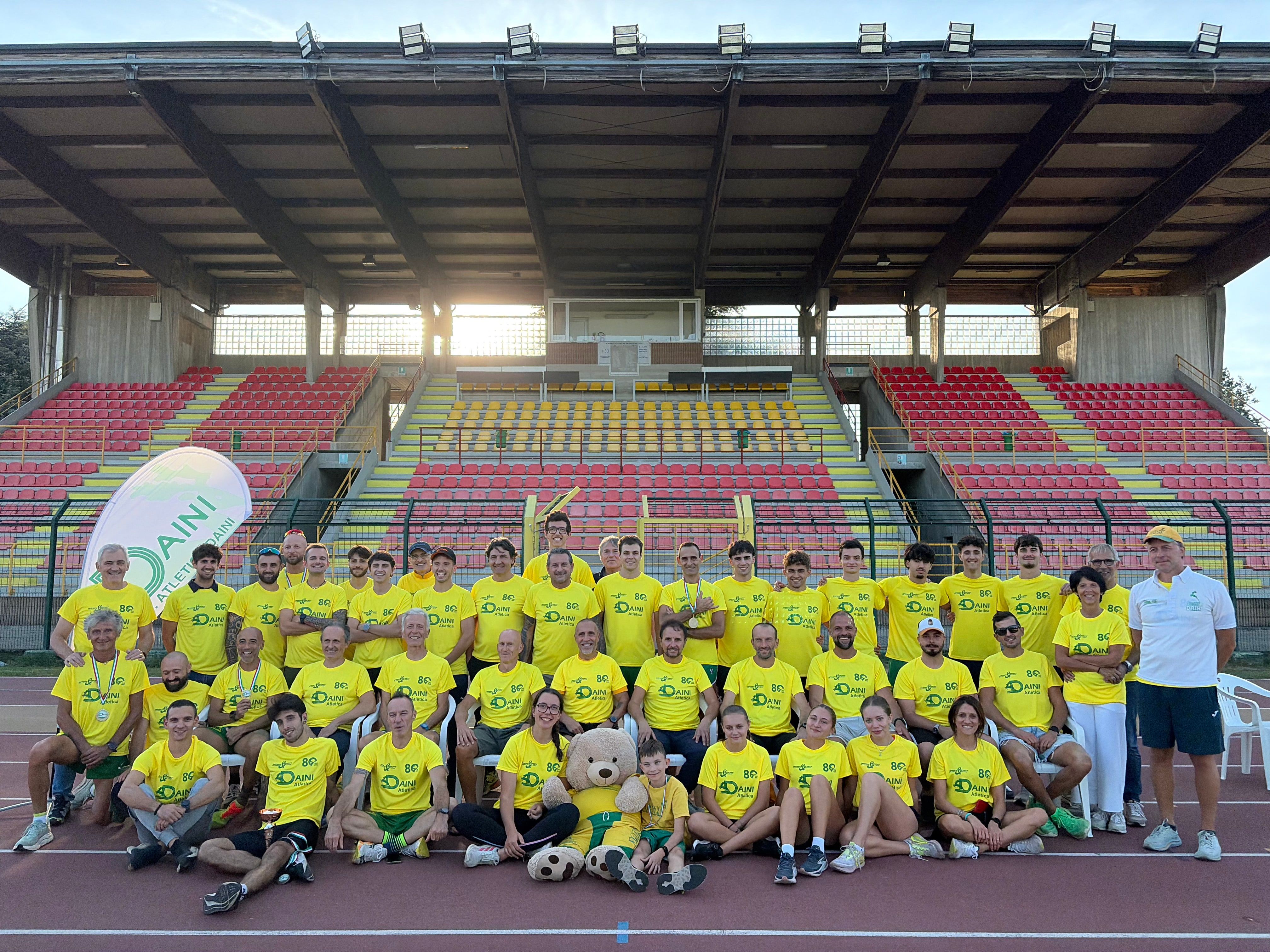 Foto di gruppo di atleti e allenatori dei Daini in maglia gialla, sulla pista davanti alle tribune