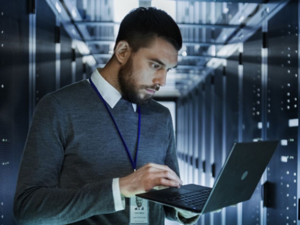 Photo of a man standing among server racks and holding a laptop