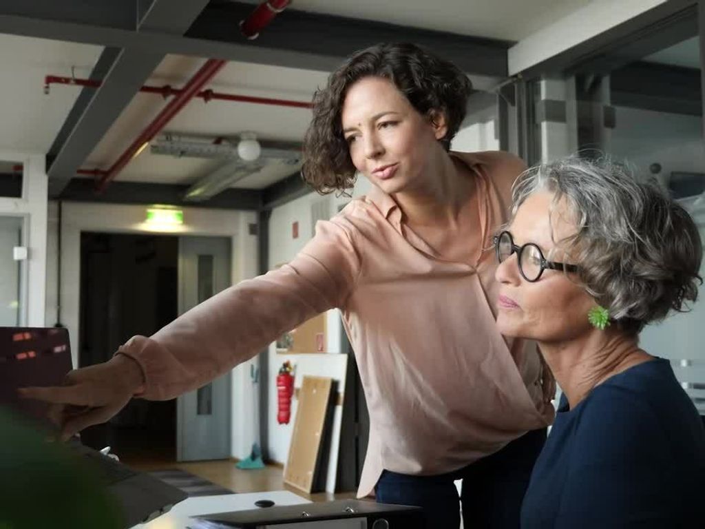 photo of two women in an office, looking at a computer screen. One is seated while the other stands next to her and is pointing at the screen.