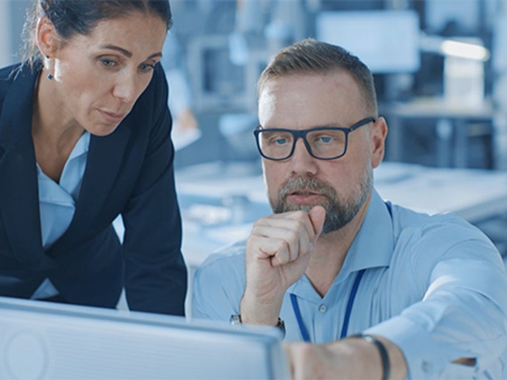 Photo of a man and a woman in an office. The man is seated and pointing at a computer screen in the foreground, while the woman is looking at the same screen from over his shoulder.