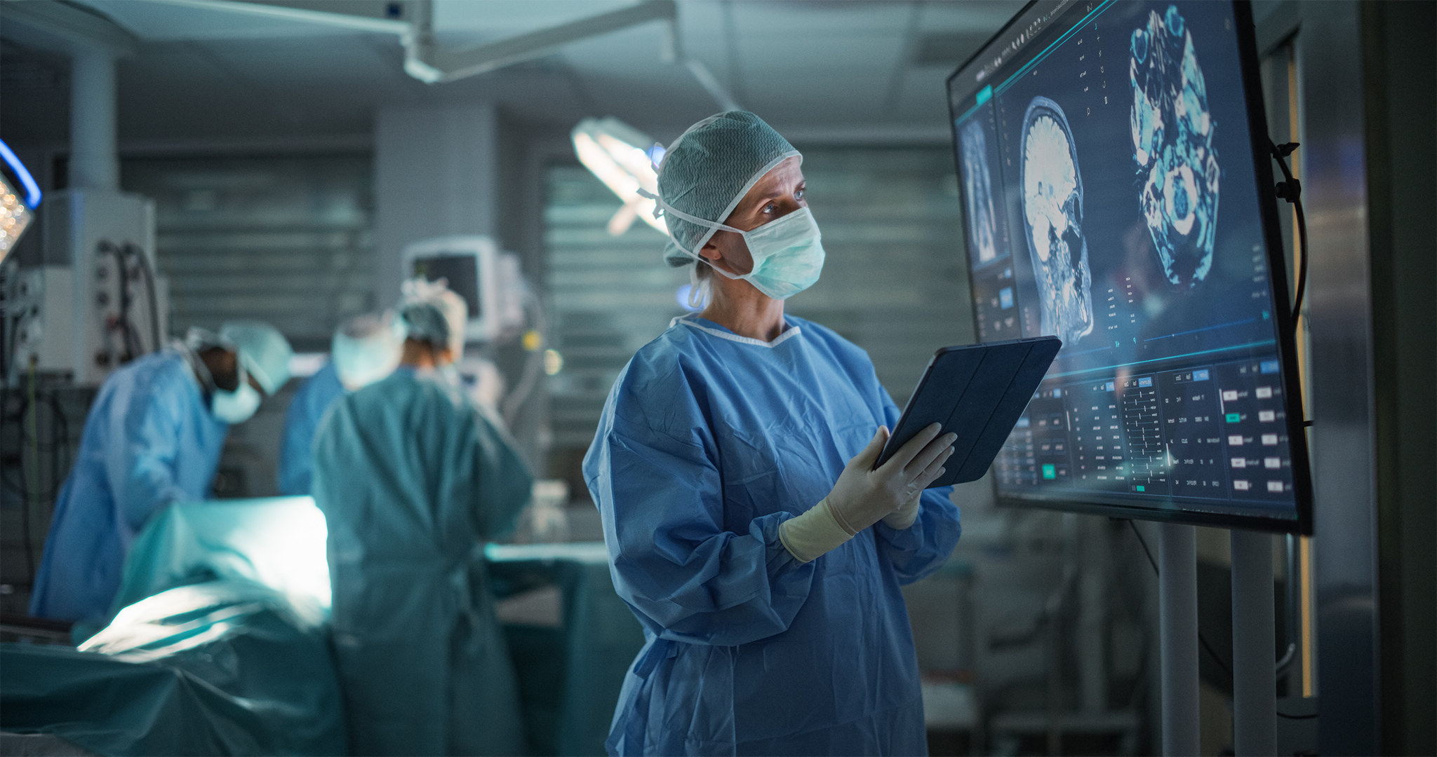 A masked medical professional in scrubs holds a tablet and is looking at a screen which has a brain scan on it