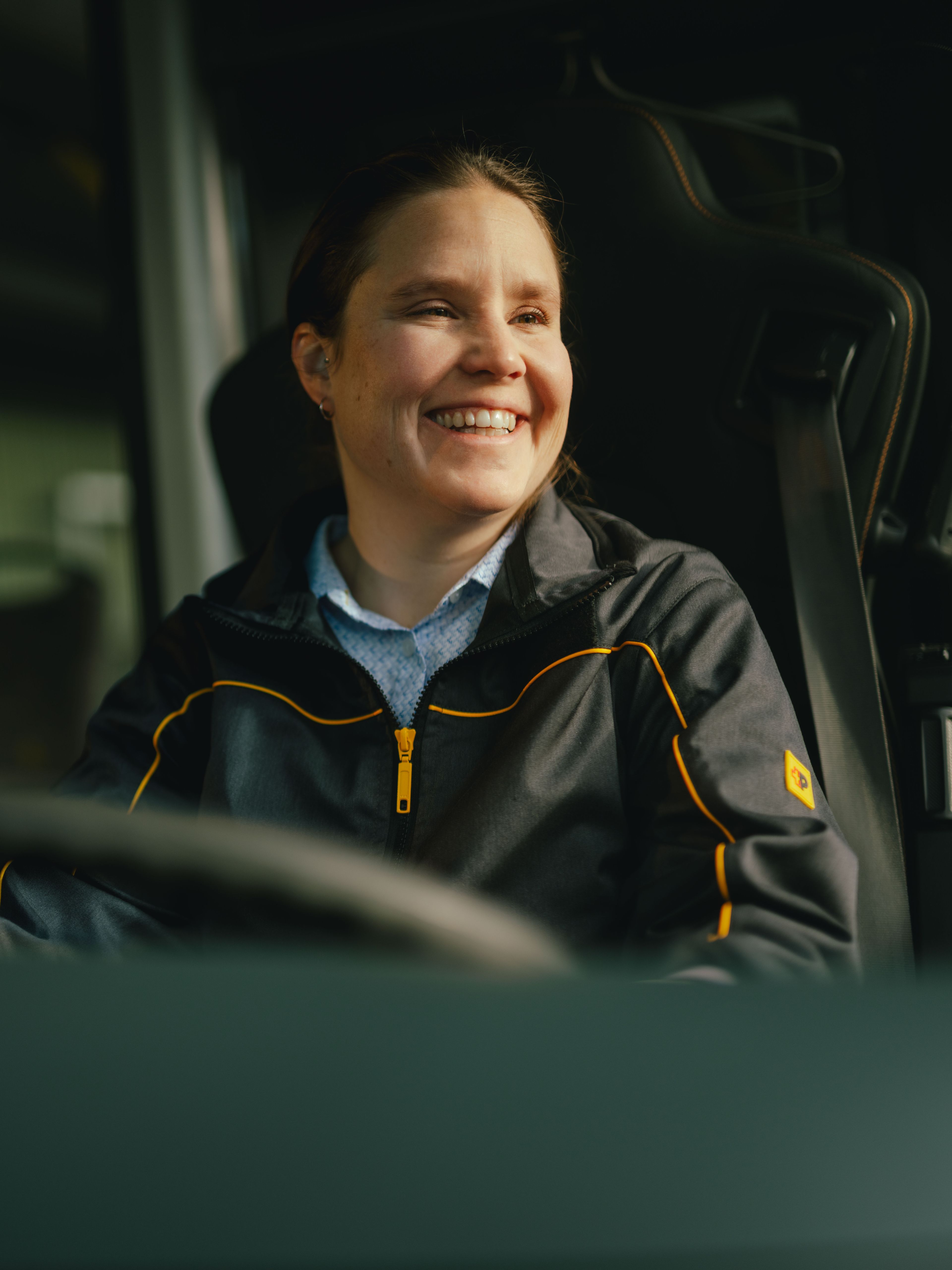Une personne est assise au volant d’un véhicule, portant une veste foncée avec des accents jaunes. L’intérieur du véhicule, y compris le volant et des poignées, est visible.
