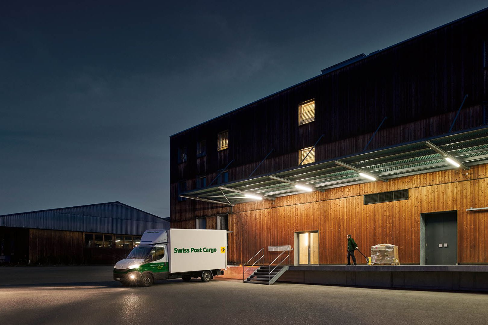 A Swiss Post Cargo night courier delivers goods to a warehouse building before dawn.