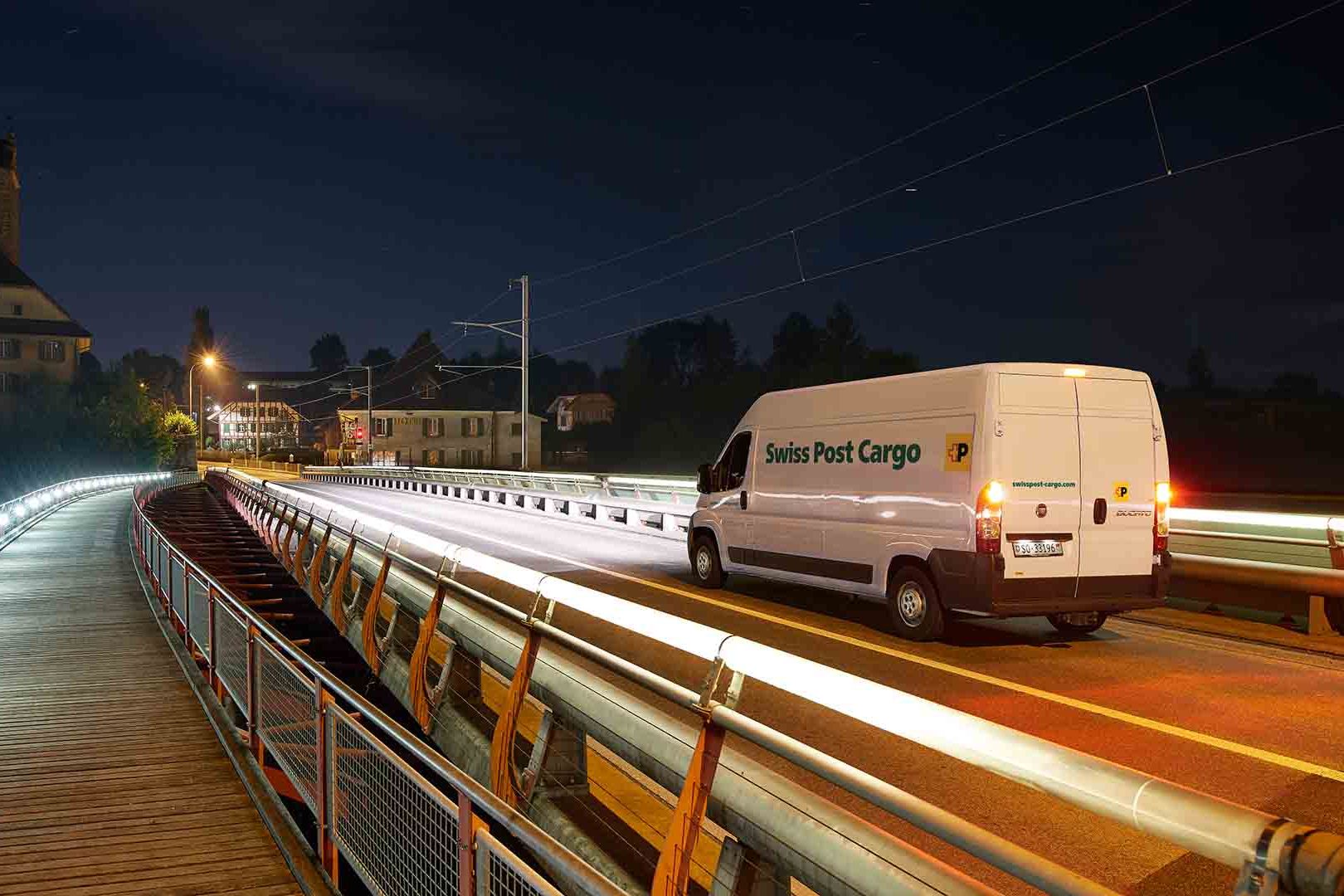 A Swiss Post Cargo delivery vehicle drives over a bridge at night with an express delivery.