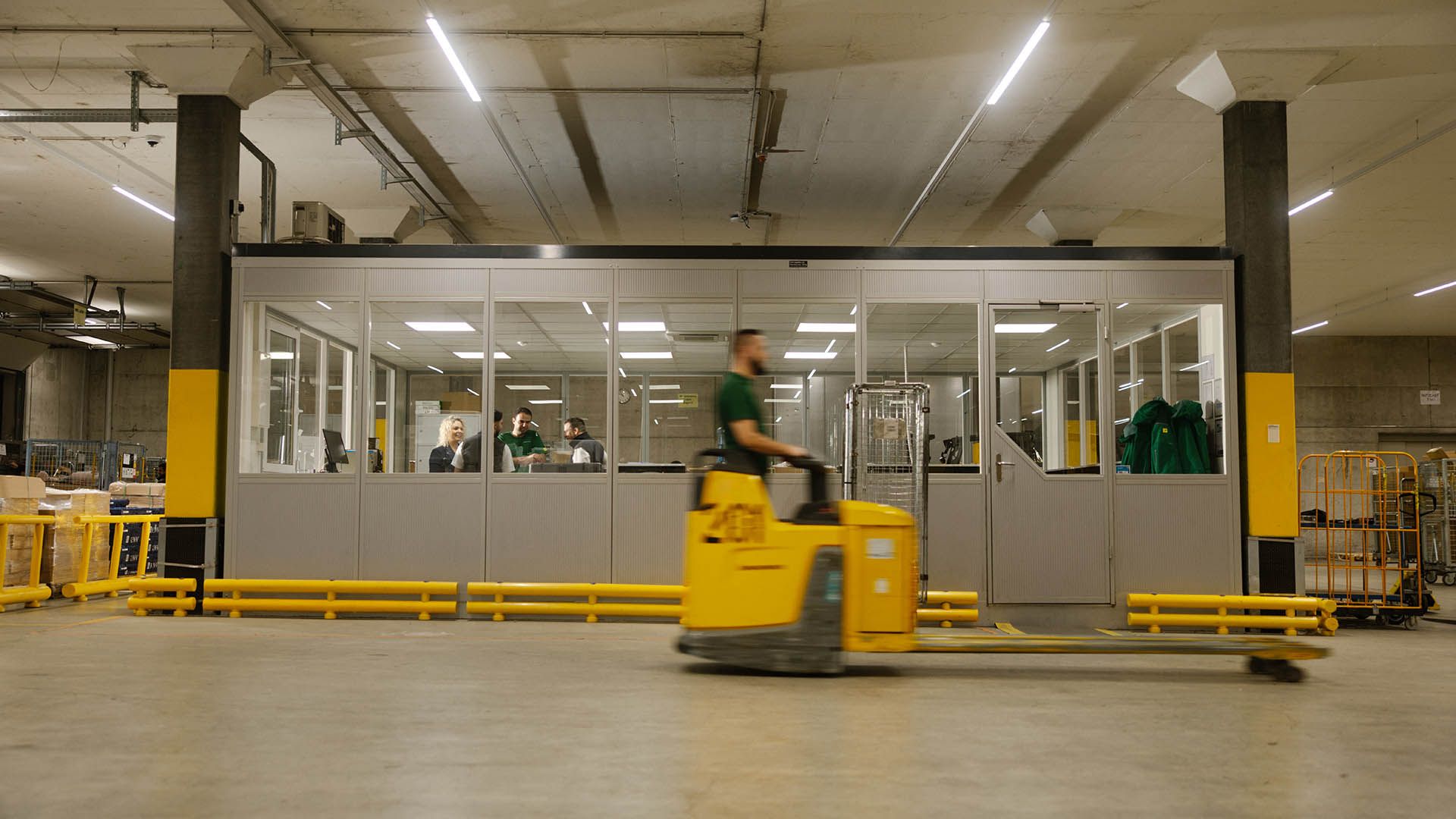 In a warehouse, a Swiss Post Cargo employee drives a pallet jack past a glassed-in room.