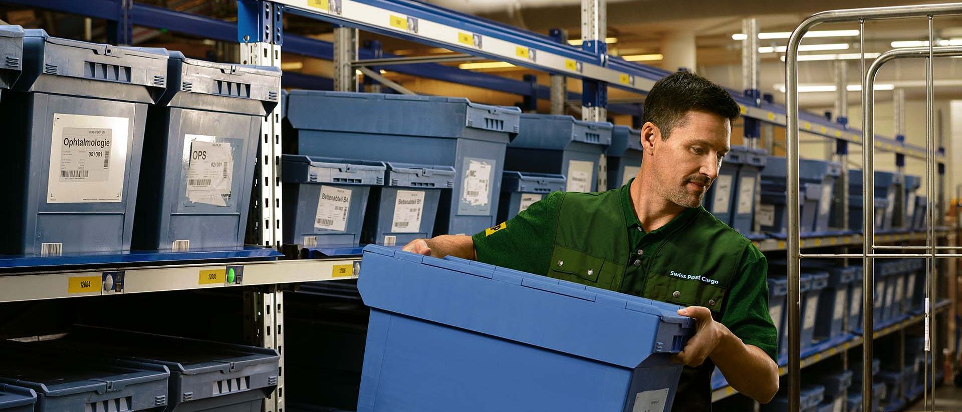 A warehouse worker loads a box with pharmaceutical goods from a shelf onto a trolley.