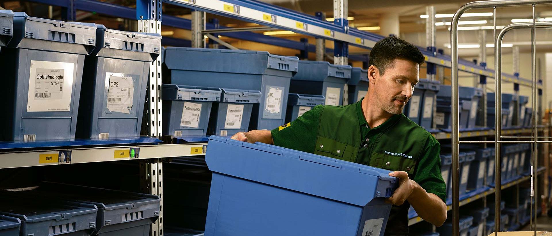 A warehouse worker loads a box with pharmaceutical goods from a shelf onto a trolley.