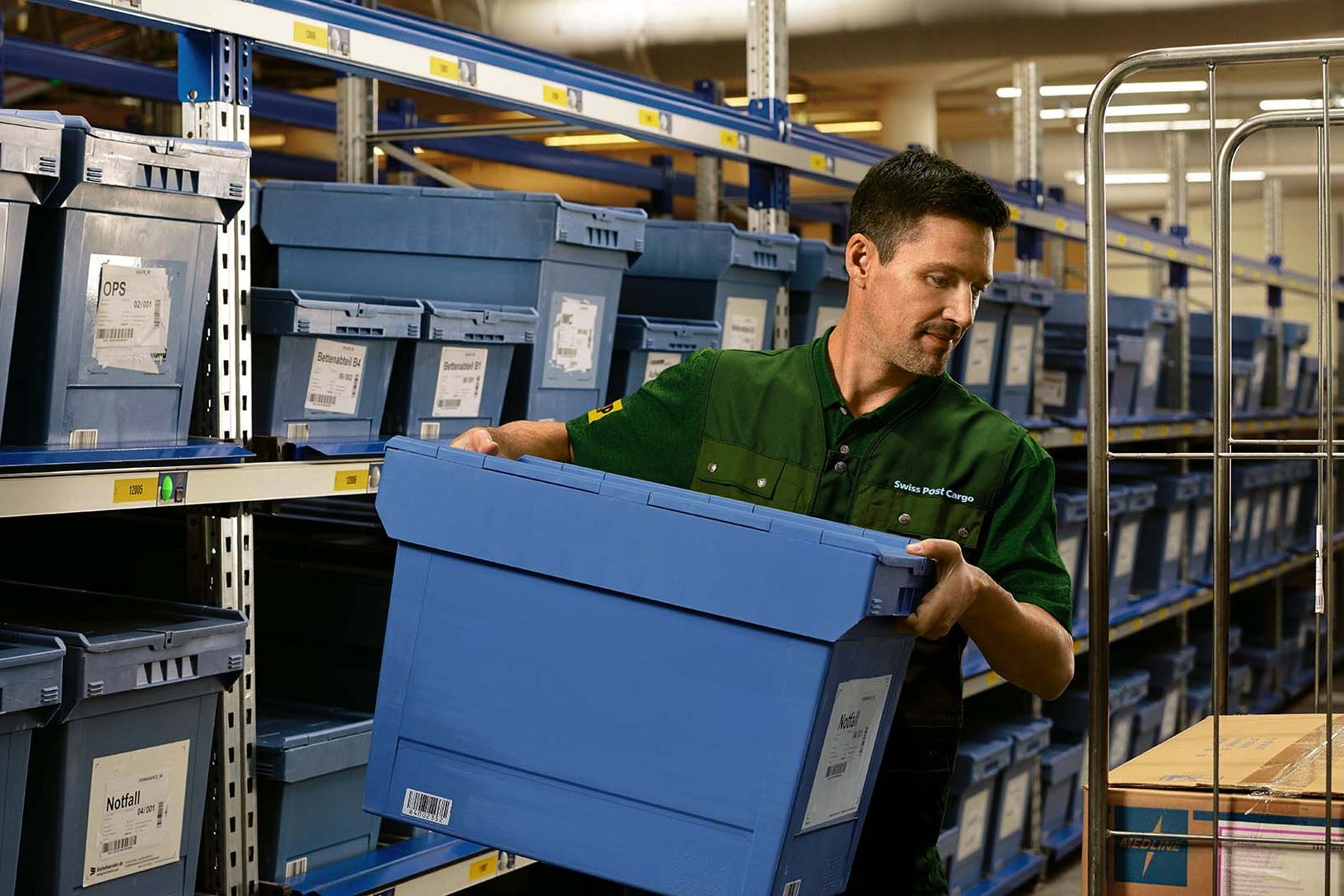 A warehouse worker loads a box with pharmaceutical goods from a shelf onto a trolley.