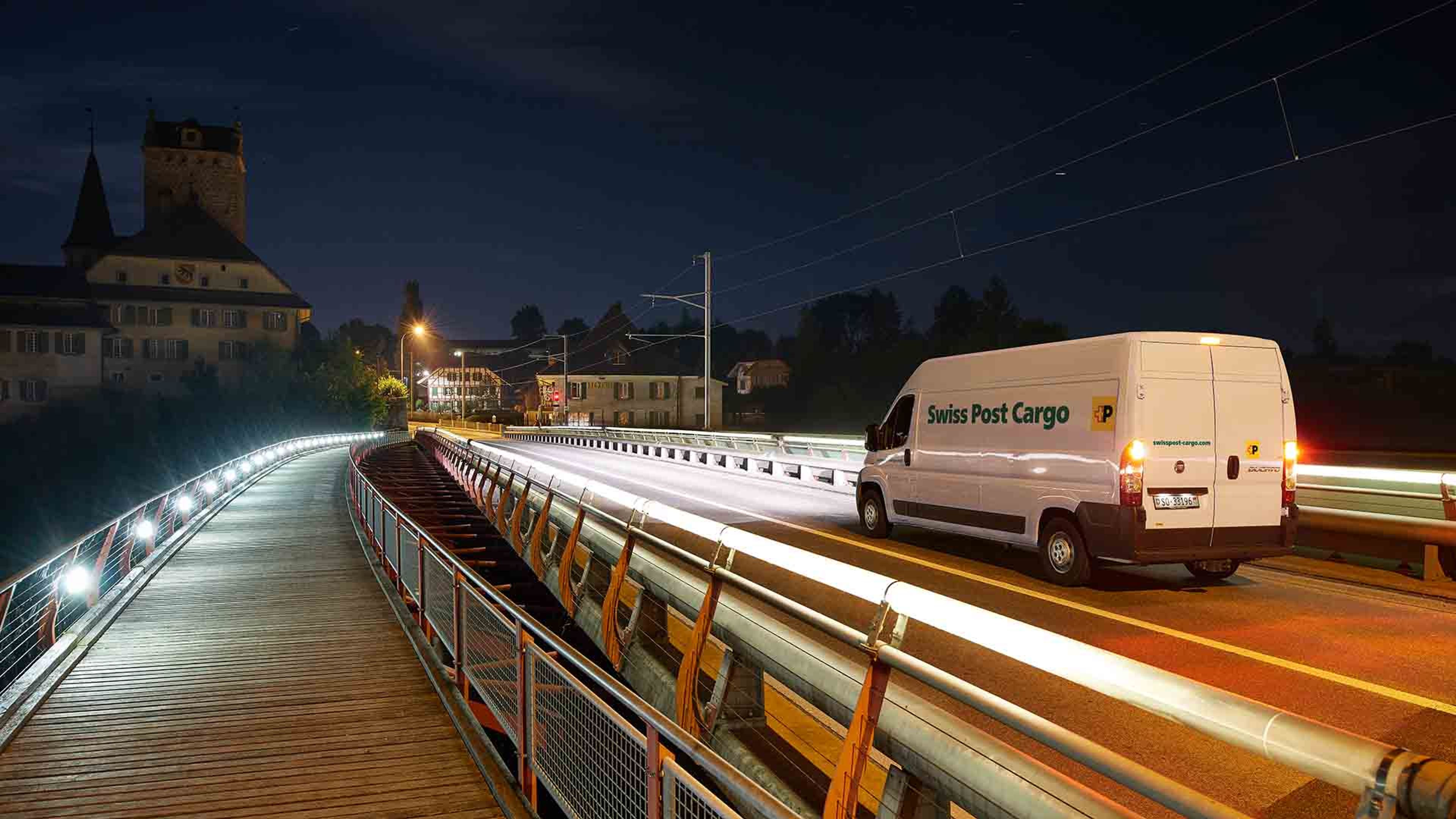 A Swiss Post Cargo delivery vehicle drives over a bridge at night with an express delivery.